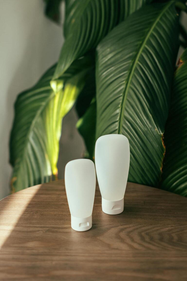 Two white cosmetic containers on a wooden table with green leaves background.